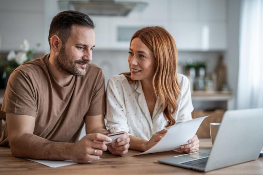 Couple reviewing household finances at kitchen table with laptop, credit card, and paperwork while planning their budget.